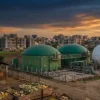 Biogas from organic waste in India: green anaerobic digester tanks and a gas-holder sphere beside a cowshed and crates of vegetable waste